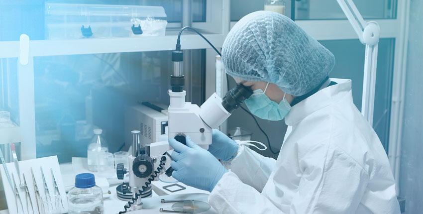 Photo of a female scientist in white lab coat peering into a microscope at a lab bench
