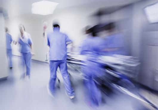 Motion blurred image of a group of people in blue hospital scrubs standing, walking, pushing a stretcher around a bend in a brightly lit corridor.