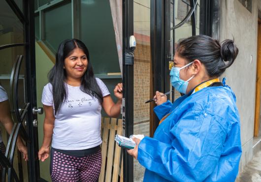 A woman in a t-shirt opens a door to talk with a woman in medical mask and protective robe.