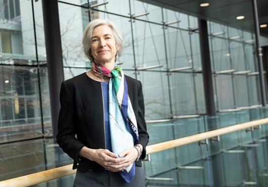 portrait photo of Jennifer Doudna, a slightly smiling white woman with chin-length gray hair, leaning against a railing in front of a wall of windows. she wears a blazer and colorful silk scarf