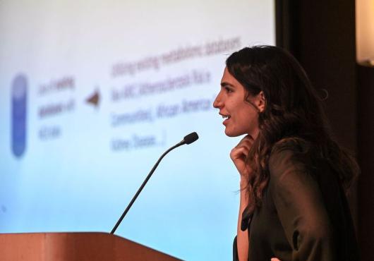 A young woman seen from the side presents at a lectern in front of a projected slide show