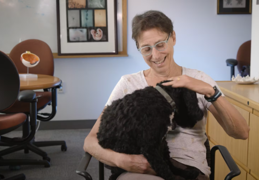 Screen shot of a woman with short hair smiling down at a cockapoo dog sitting in her lap. She is in an office with a table and chairs and framed art in the background.