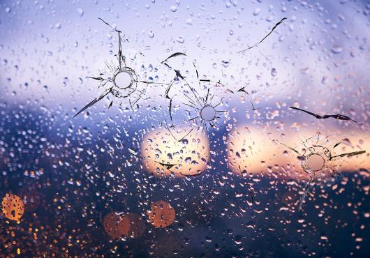 City lights reflected in a raindrop-speckled window, the glass shattered by several round bullet holes with jagged cracks.