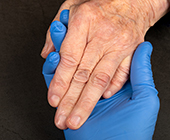 A hand in a blue latex glove holds the ungloved hand of an older white person