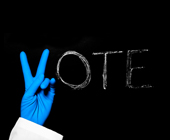 Photo of chalkboard with the word vote - the V is being made by the hand of a person with surgical gloves on.