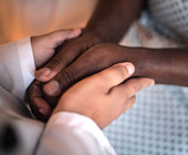 Close up of a white coated doctor's hands holding the hands of a black patient wearing a hospital Johnny.
