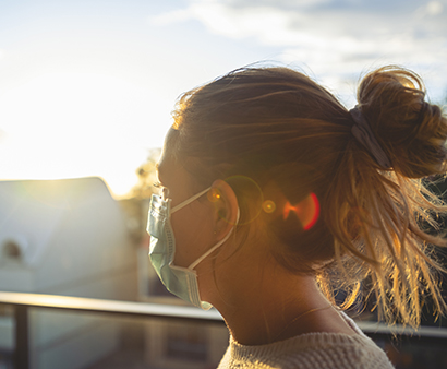 Woman wearing a face mask looking from her balcony at sunset. She is alone and looks a little sad or depressed. 