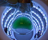 A flask of green liquid held by a gloved hand is silhouetted at the top of a cylinder with rings of lights