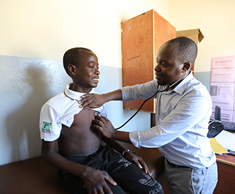 A teenage boy sits on an examination table in a clinic as a man listens to his heart with a stethoscope.