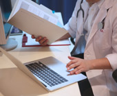Photo of physician holding a pen and a notebook and typing on a computer keyboard