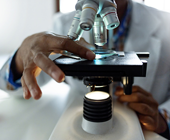 Close up of person in a white coat with black hands holding a microscope