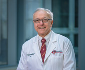 Photo portrait of Dean Daley smiling. He's wearing a white coat with Harvard Medical School logo and HMS crimson tie and wearing glasses