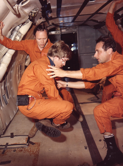 three men in orange flight suits functioning in a weightless environment