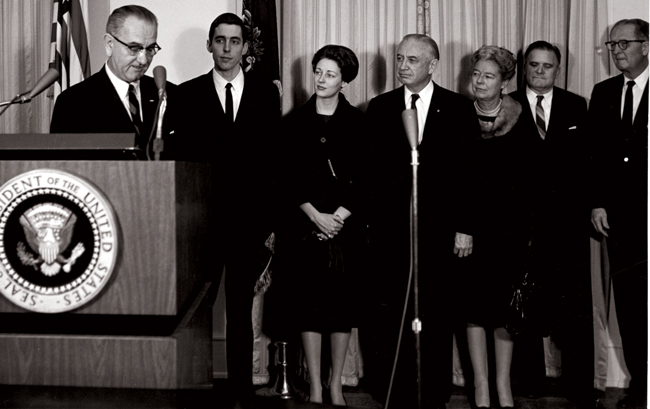 archival photo showing President Lyndon Johnson at a podium with a number of people standing to the right of him