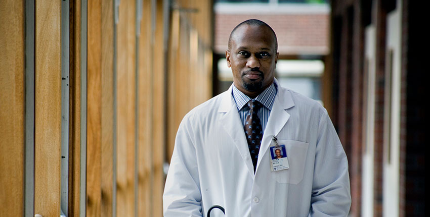 Stultz in a white coat with stethoscopes and hands in his pockets standing in a hallway