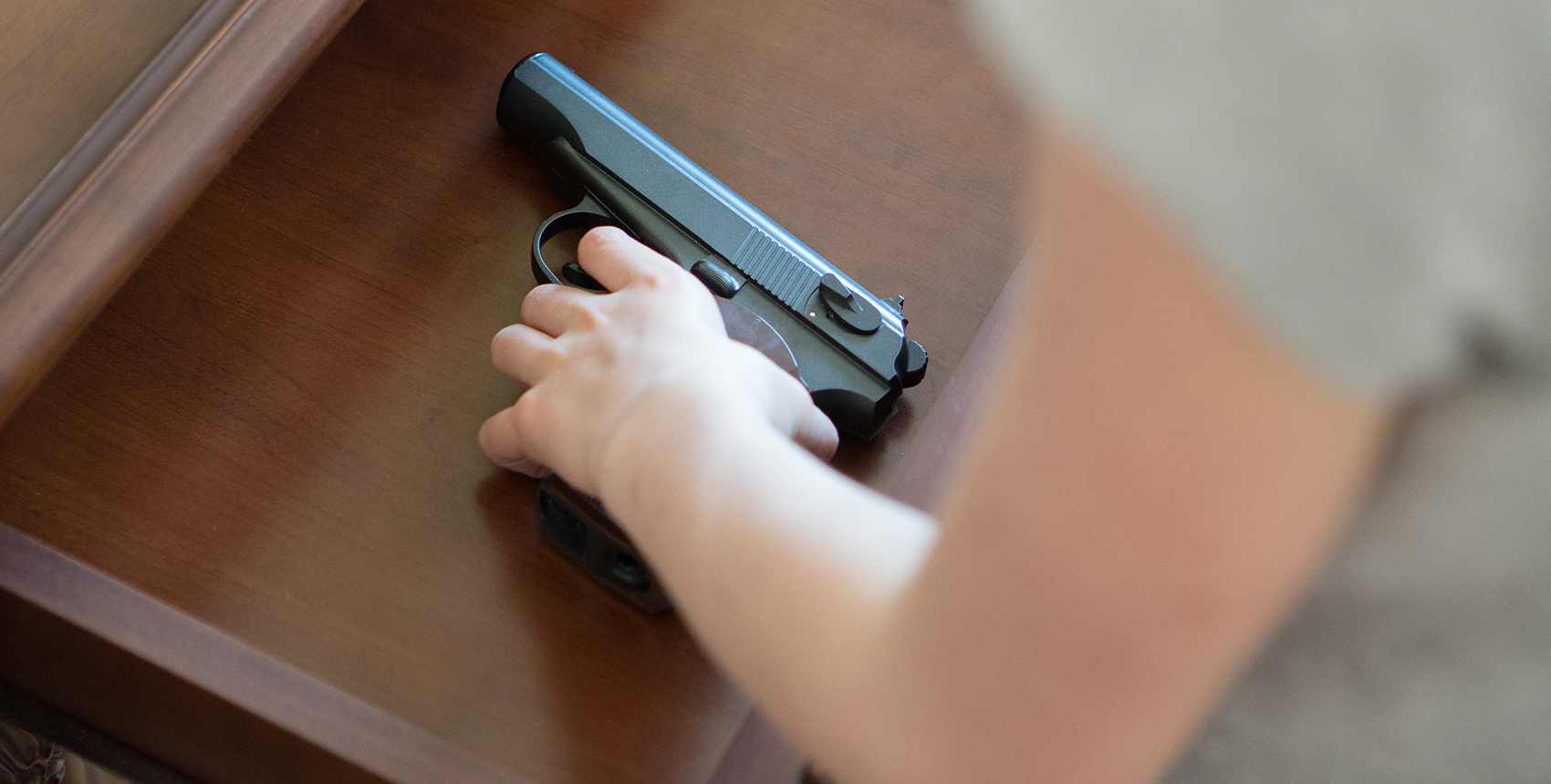 A child's hand reaches into an open drawer in a wooden dresser to pick up a black handgun with a brown handle.