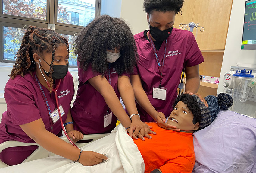 Three high school students in scrubs and masks work on a medical mannequin, one has a stethoscope, one is doing chest compressions, one is checking airway