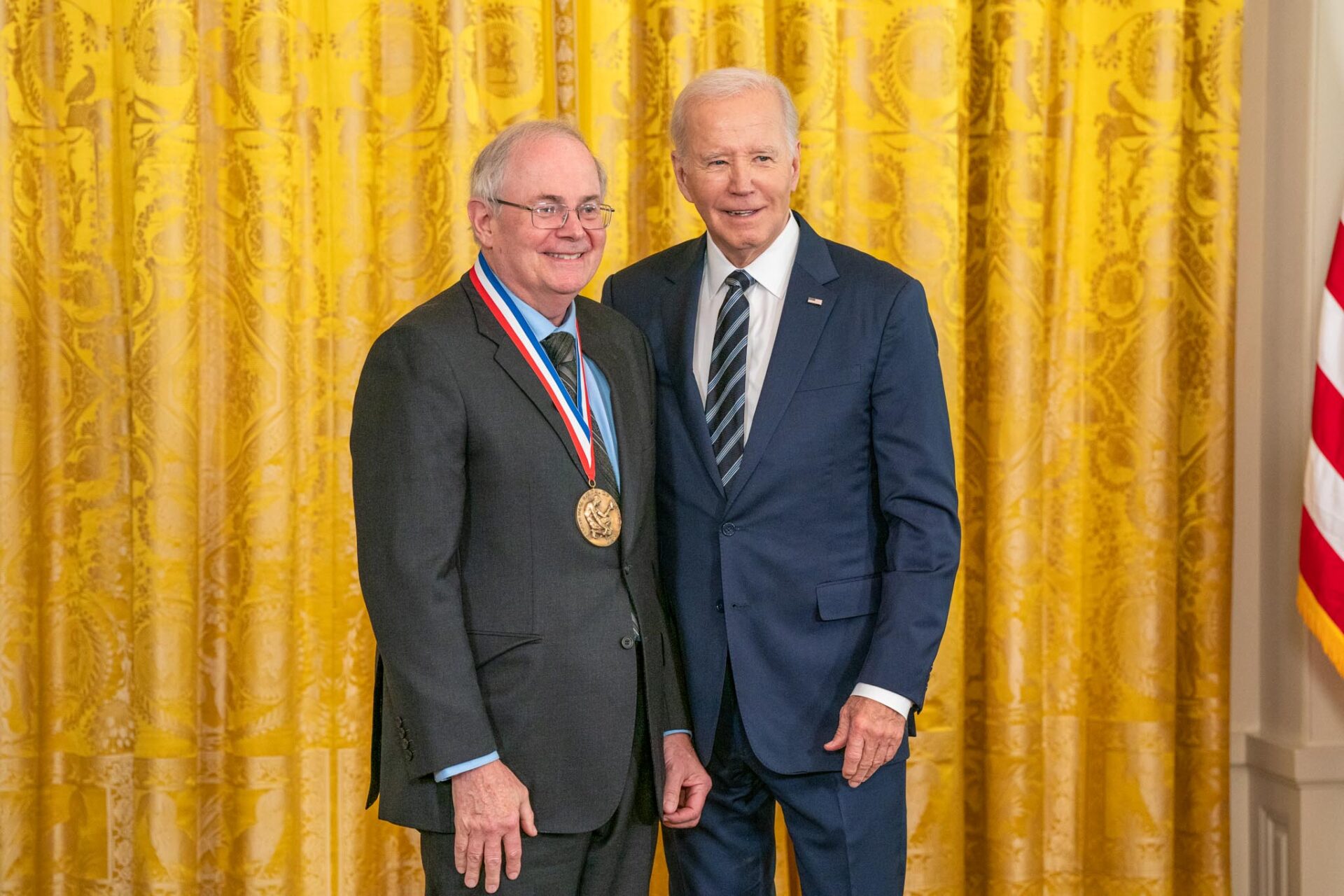 U.S. President Joseph Biden stands in a suit beside Gregory Petsko, a gray-haired white man also in a suit. Petsko wears his new National Medal of Science. Both are smiling. They stand before a gold curtain.