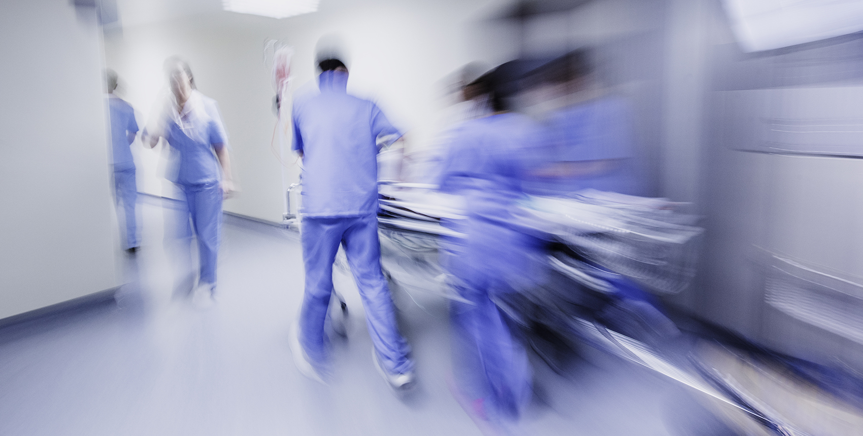 Alt text: Motion blurred image of a group of people in blue hospital scrubs standing, walking, pushing a stretcher around a bend in a brightly lit corridor.