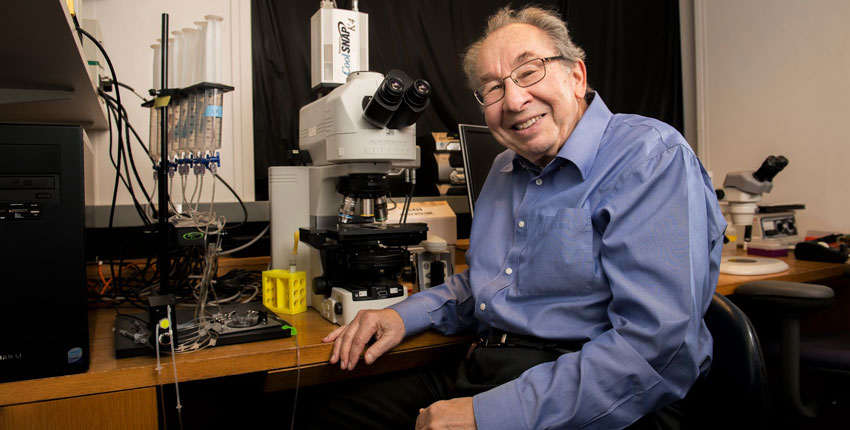 A photo of Ed Kravitz in a blue shirt sitting in front of a microscope at a lab bench