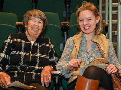 An older woman in a plaid blazer sits in auditorium seats next to a younger woman in a tan vest. Both are smiling.