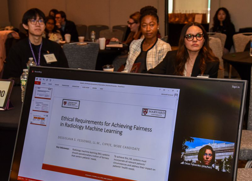 a laptop screen shows a woman presenter and a slide show. in the background, attendees with name tags look on