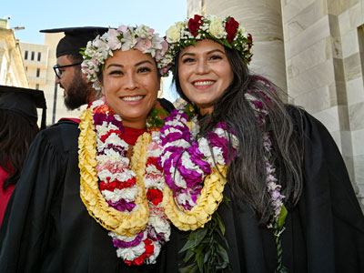 Two people in graduation robes and flower leis posing side by side