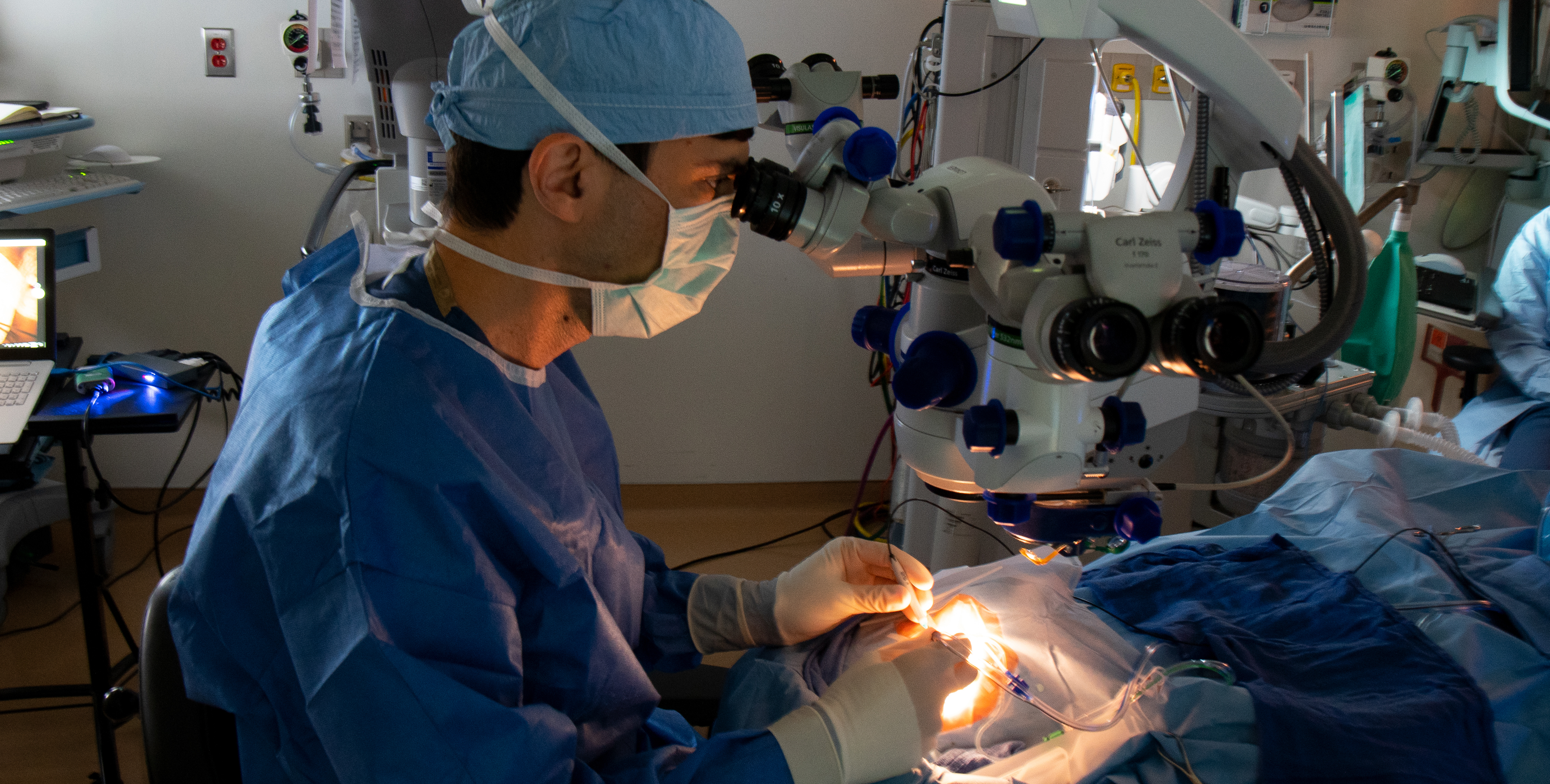 Wearing tanslucent white gloves and a blue cap, mask, and medical garb, a surgreon manipulates several objects connected to wires and tubes above the patient's eye. The patient's face is invisible in the intense glow of the operating lights.