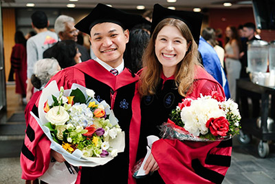 Photo of students wearing graduation caps and gowns, holding bouquets of flowers.