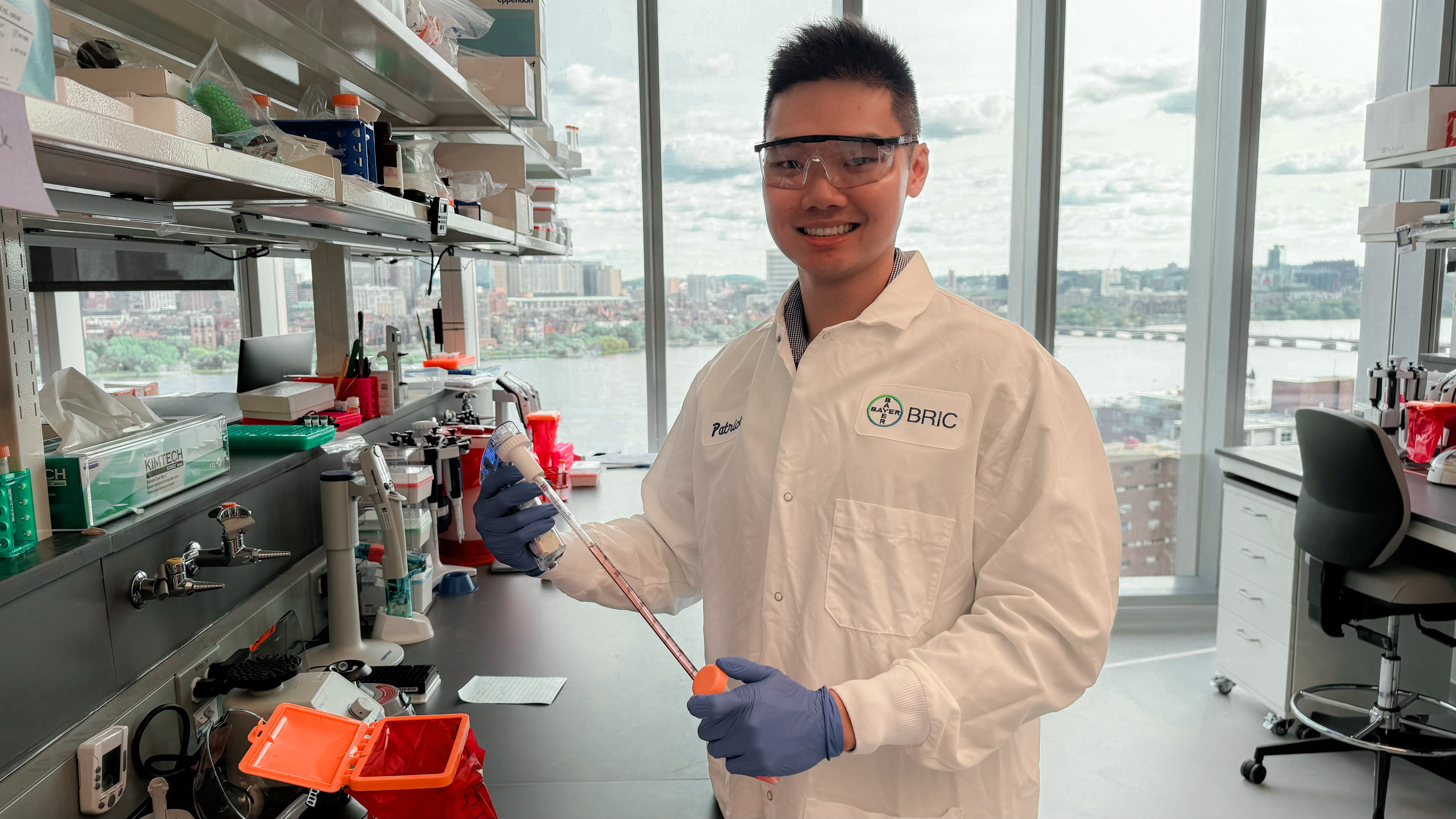 A young man in safety glasses, lab coat, and gloves holds a pipetting instrument in a lab. Behind him is a wall of windows overlooking Boston.