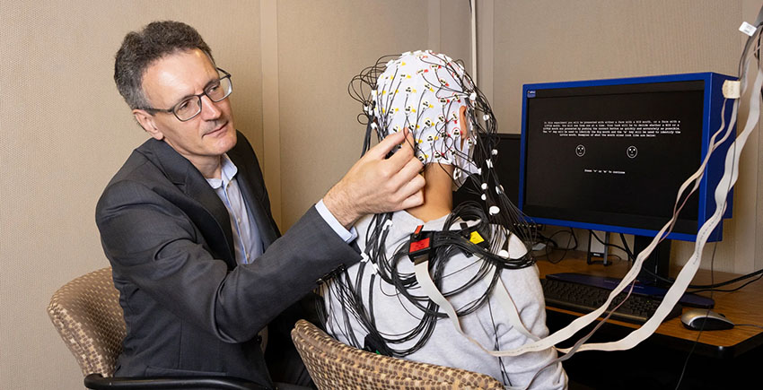 A man adjusts an electrode-filled cap on the head of a patient whose back is to the camera. They sit in front of a computer.