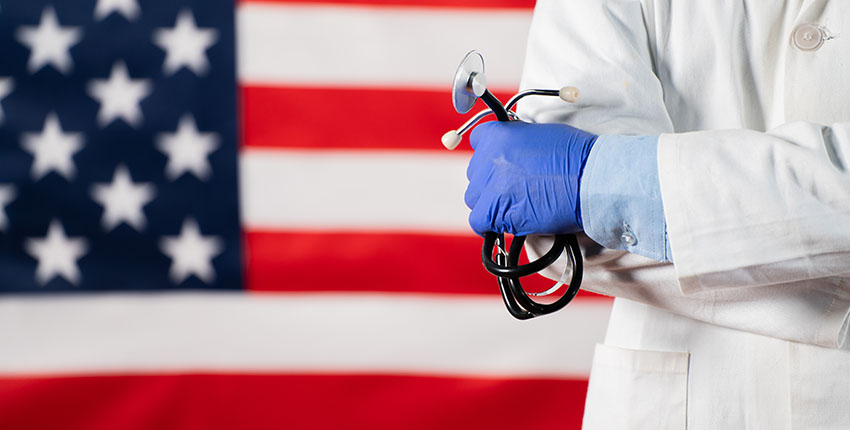 A health care practitioner wearing a white coat and holding a stethoscope stands in front of an American flag