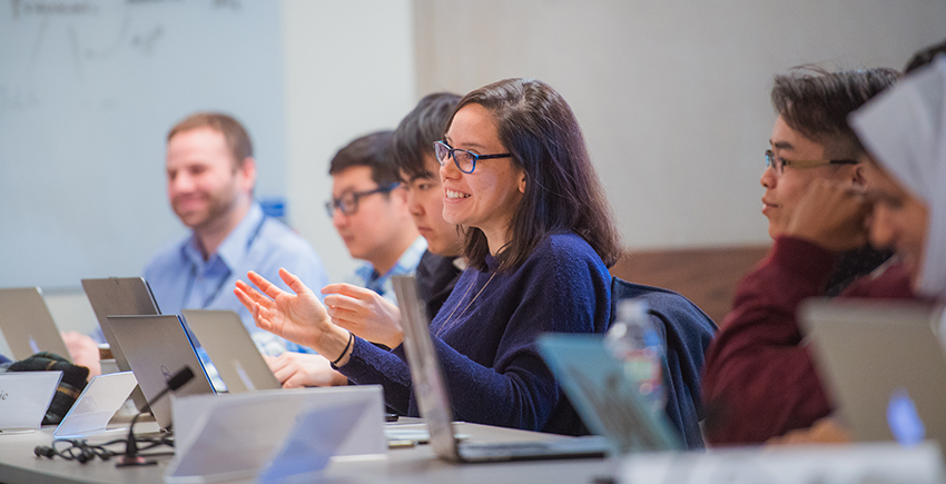 Six trainees at a conference table with laptops in front of them. The one at center speaks with a smile and gesturing hands.