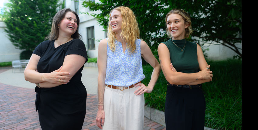 Three young women stand in courtyard smiling at each other