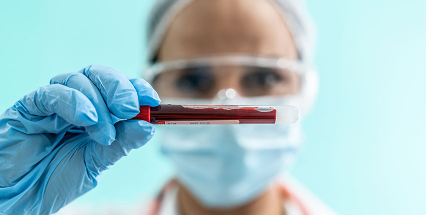 A person wearing protective gloves, glasses, and mask holds a small vial containing a blood sample.