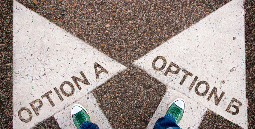 An aerial view of two feet in green sneakers, one standing on a large white arrow labeled “option A” and the other standing on a large white arrow labeled “option B”