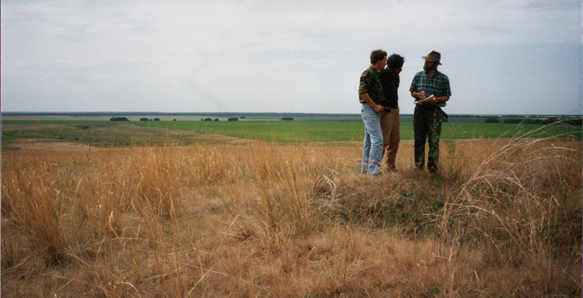 Three people stand on a hill overlooking a grassy plain