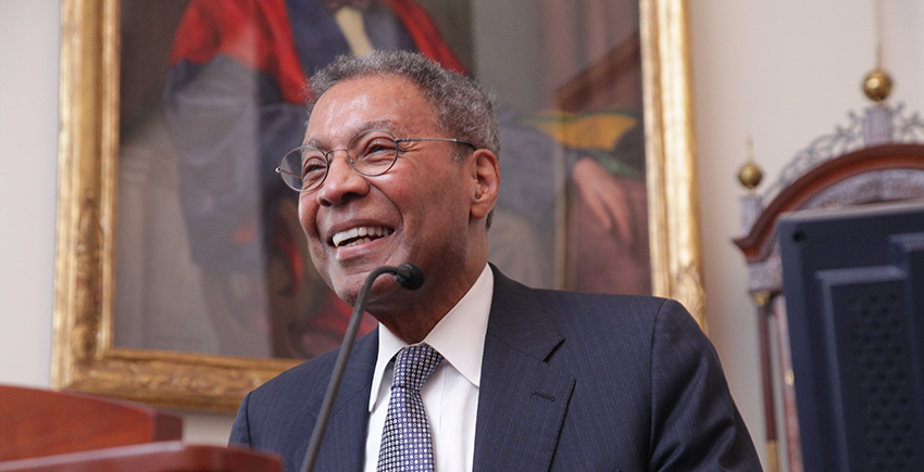 A man in a suit smiles at a lectern in front of a painting of someone in academic robes