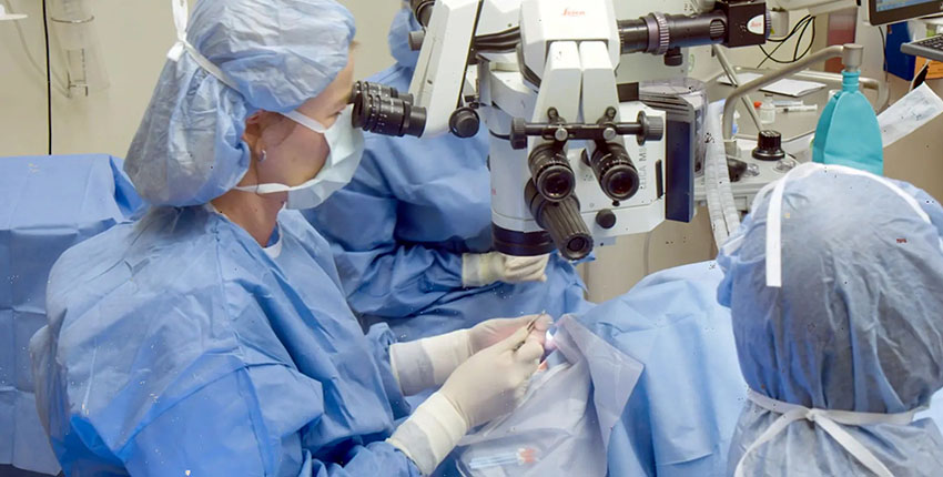 In an operating room, three people in blue surgical gowns, masks, and head covers look through microscopes while performing surgery on a patient.