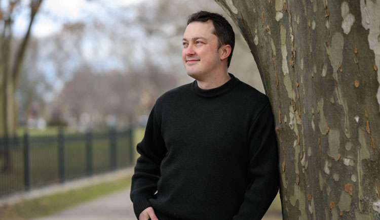 A man leans against a tree, smiling and looking off into the distance