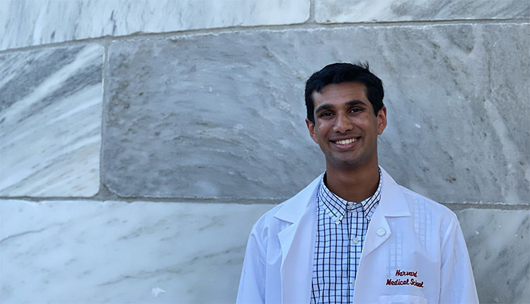 A man in a white coat stands in front of a marble wall that says Harvard Medical School