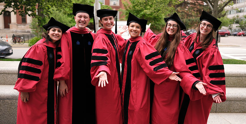 Five women and one man in academic regalia smile and stretch out their hands, tah dah.