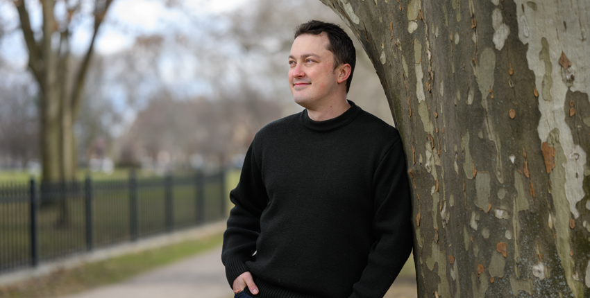 A man leans against a tree, smiling and looking off into the distance