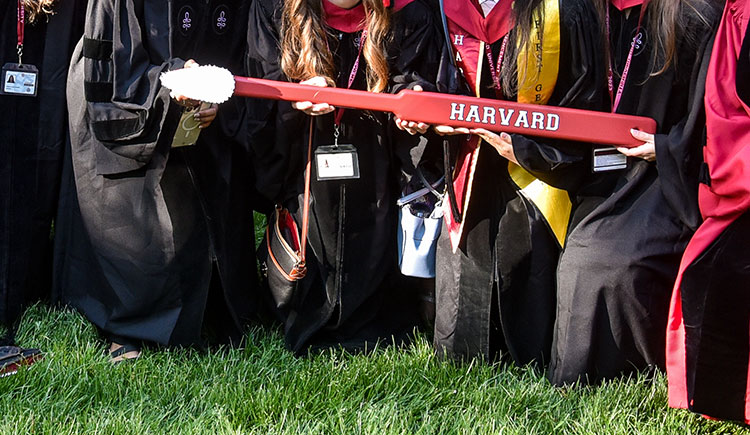 A giant toothbrush embossed Harvard held by grads in regalia