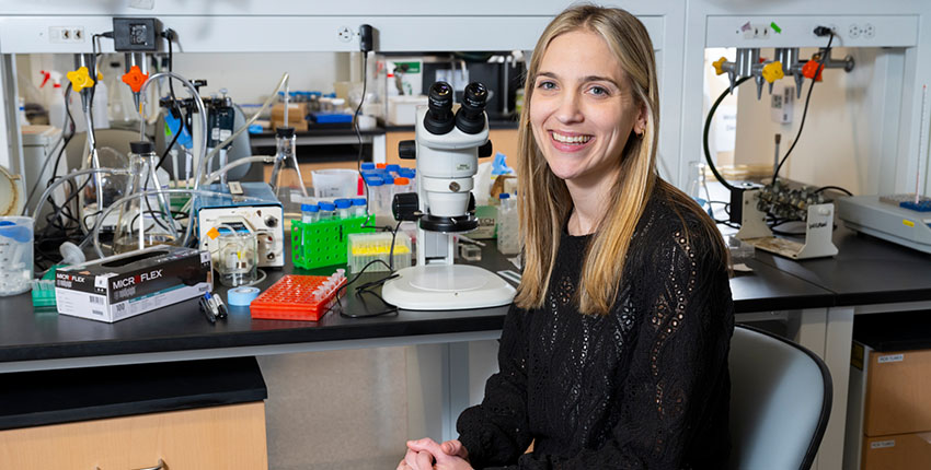 A researcher sits on a chair next to a microscope in a lab space