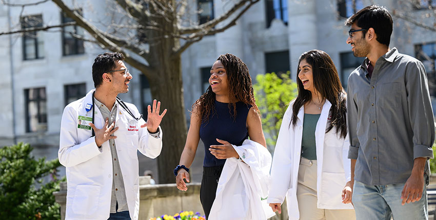 Four medical students, two in white coats, smiling and walking together on a sunny day with a marble columned building in background.