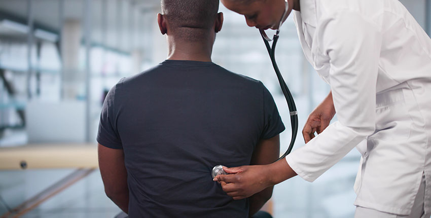 A doctor in a white lab coat uses a stethoscope to listen to the back of a seated patient wearing a dark t-shirt, in a bright, modern medical office.