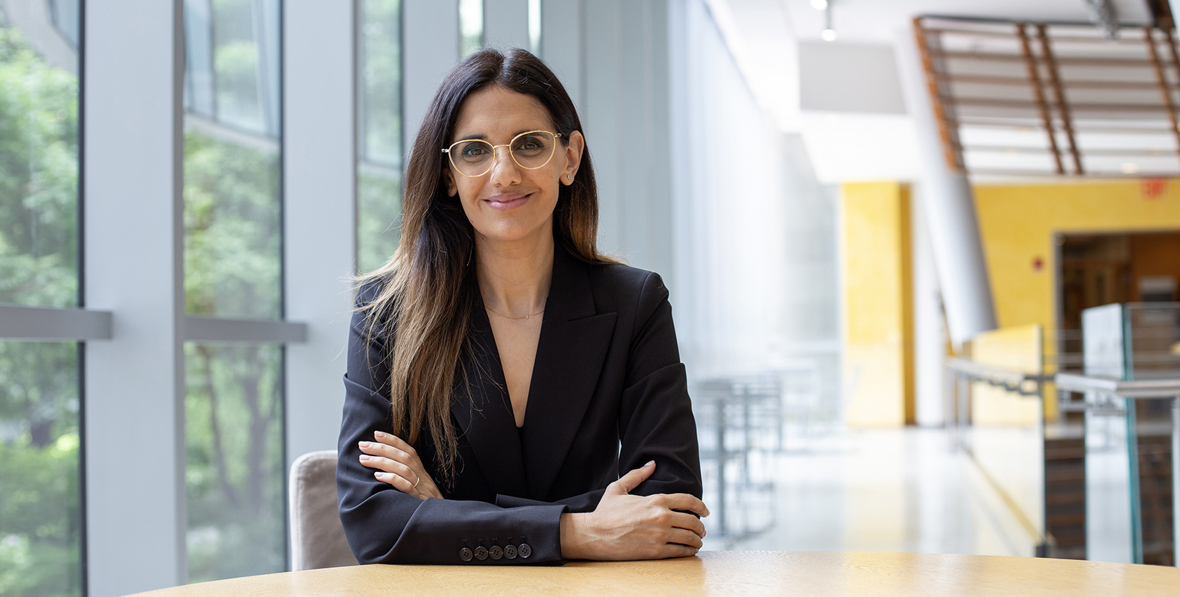 A woman with long, straight hair and glasses sits at a round wooden table in a modern, sunlit building with large windows. She is wearing a black blazer and smiling confidently with arms crossed.