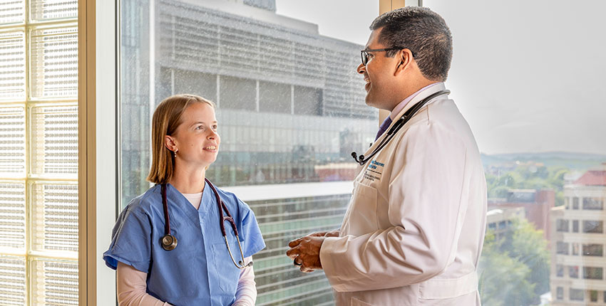 A woman in blue scrubs and a man in a white coat, both wearing stethoscopes, stand talking to each other in front of a window.