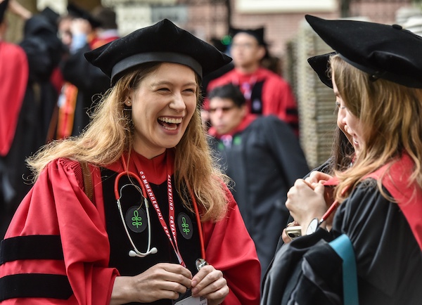 A MD-PhD student dressed in graduation regalia greets other colleagues with a wide smile.